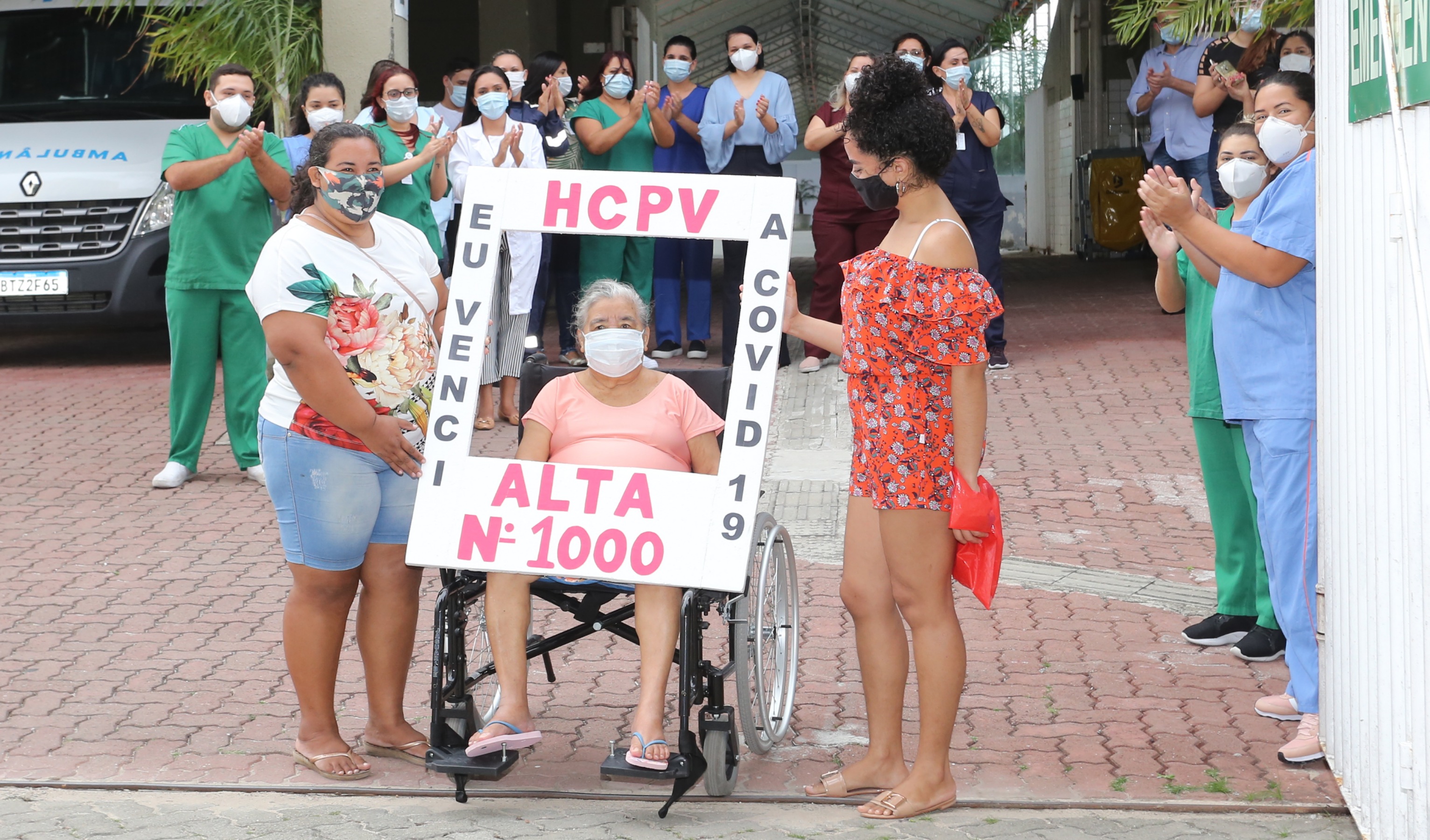 senhora em cadeira de rodas com cartaz escrito 'eu venci a covid-19. alta número 1000', profissionais de saúde aplaudindo ao fundo, rapaz e duas moças em pé ao lado dela. todos usam máscara